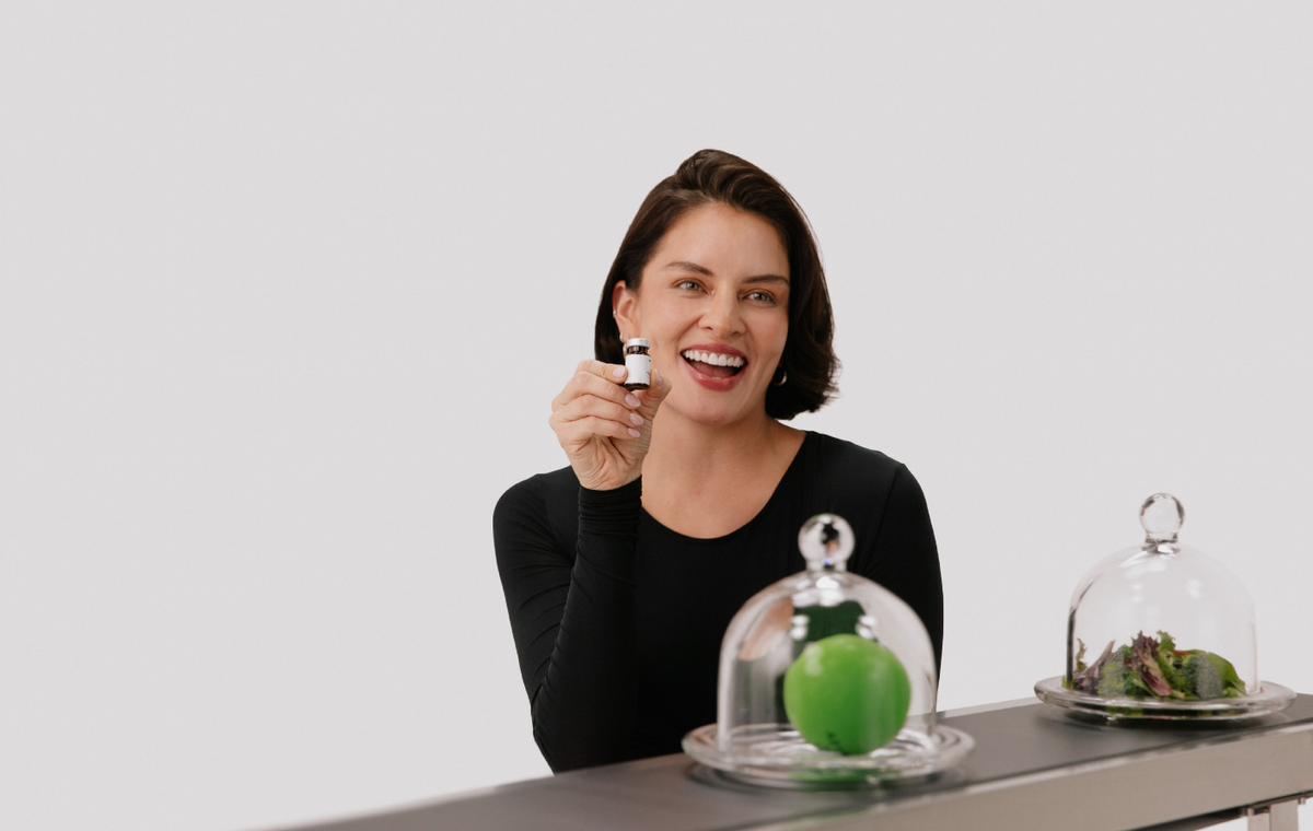 smiling woman holding a medical vial next to a cloche with a green apple in it on a gray background