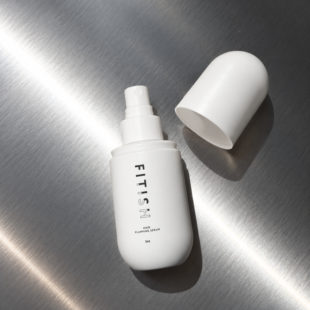 Fitish Hair Plumping Serum spray bottle with the fine mist sprayer showing, cap beside it on brushed steel