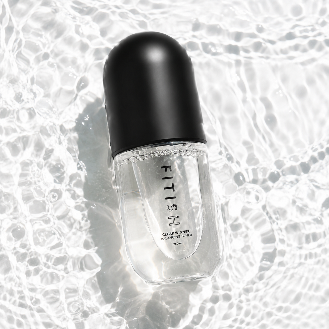 A clear cosmetic bottle with a black cap, labeled FITISH CLEAR MINIS COFFEE, placed on a glossy, wet reflective surface.
