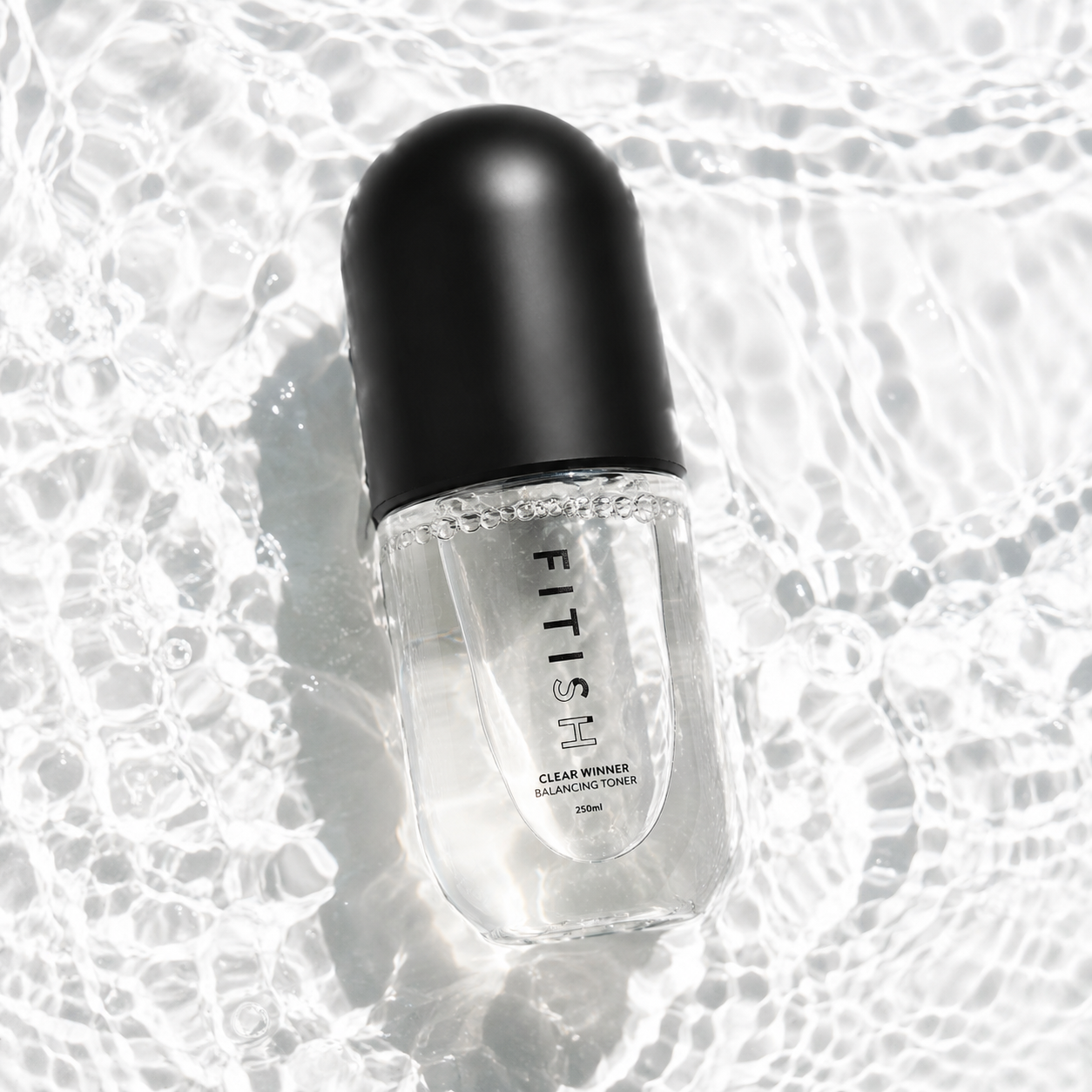 A clear cosmetic bottle with a black cap, labeled FITISH CLEAR MINIS COFFEE, placed on a glossy, wet reflective surface.